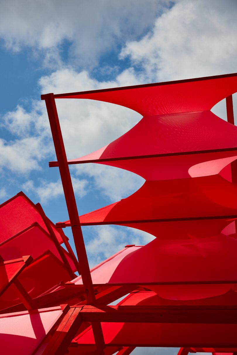 Red geometric structures made of fabric and wood are seen against a backdrop of a partly cloudy blue sky.