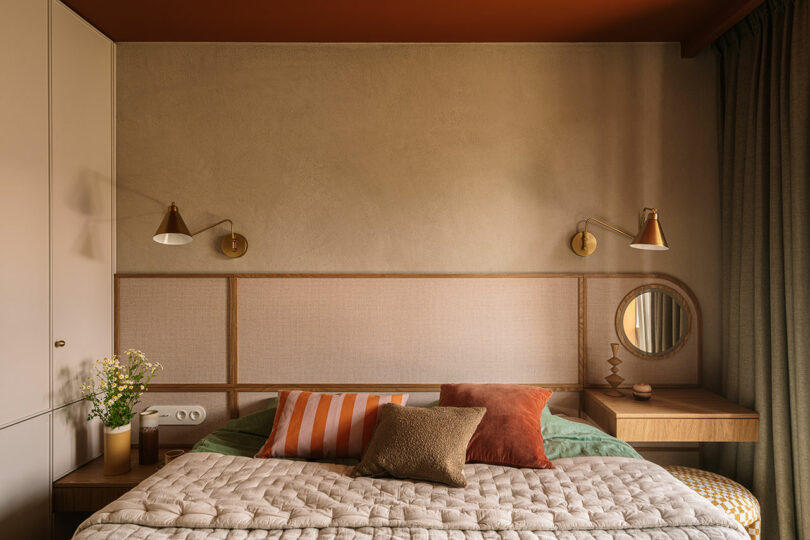 A neatly made bed with earth-toned pillows and blanket sits against a beige wall, flanked by wall lamps and built-in nightstands; a round mirror and green curtains are on the right.