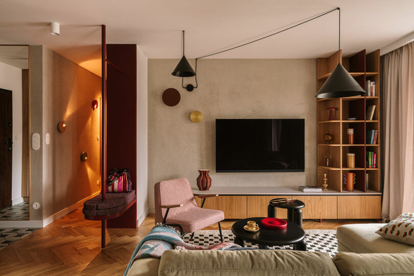 A modern living room with a wall-mounted TV, open shelving, contemporary lighting, a pink armchair, a green sofa, and geometric decor accents.