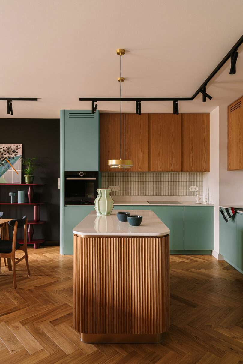 Modern kitchen with light wood and green cabinets, a marble-topped island, pendant lighting, and herringbone wood flooring.