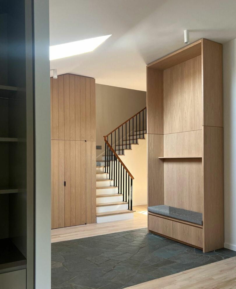 A modern entryway by Nicki Gitlin features light wood cabinetry, a built-in bench, slate flooring, and a staircase with sleek black railings leading upstairs.