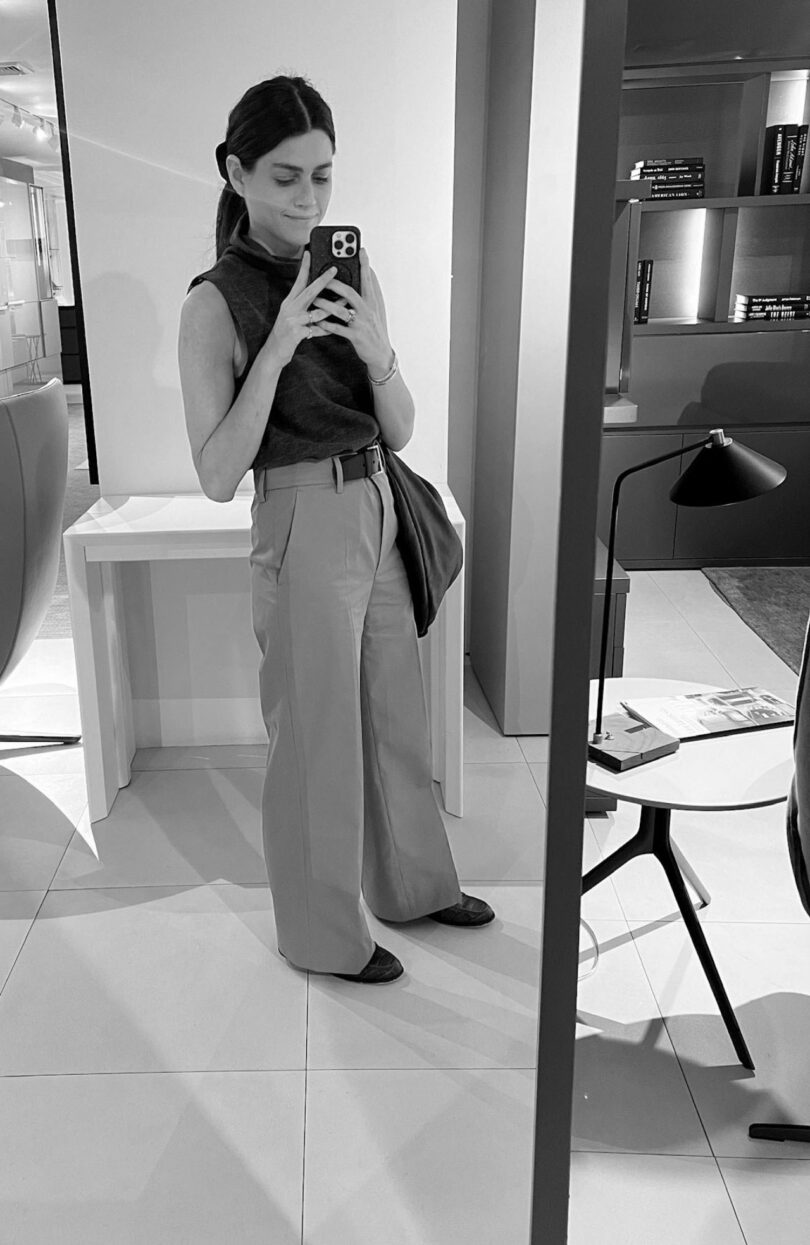 Nicki Gitlin takes a mirror selfie in a modern, minimalistic room furnished with a sleek desk, chair, lamp, and organized shelves.