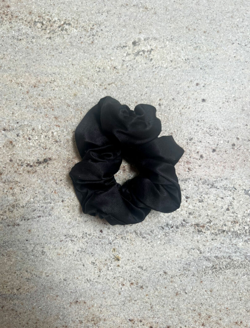 A black fabric scrunchie designed by Nicki Gitlin is lying on a light-colored, speckled surface.