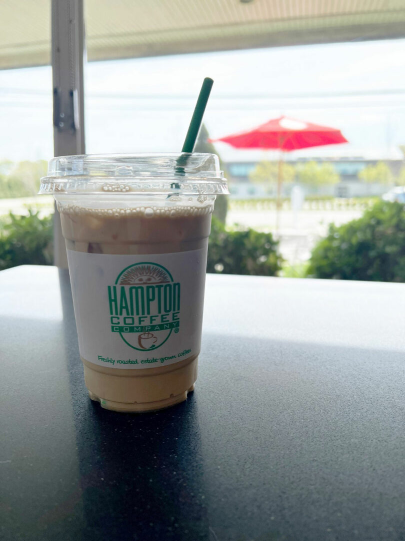 A plastic cup of iced coffee with a green straw from Hampton Coffee Company sits on a table; an outdoor scene with a red umbrella is visible in the background, captured by Nicki Gitlin.