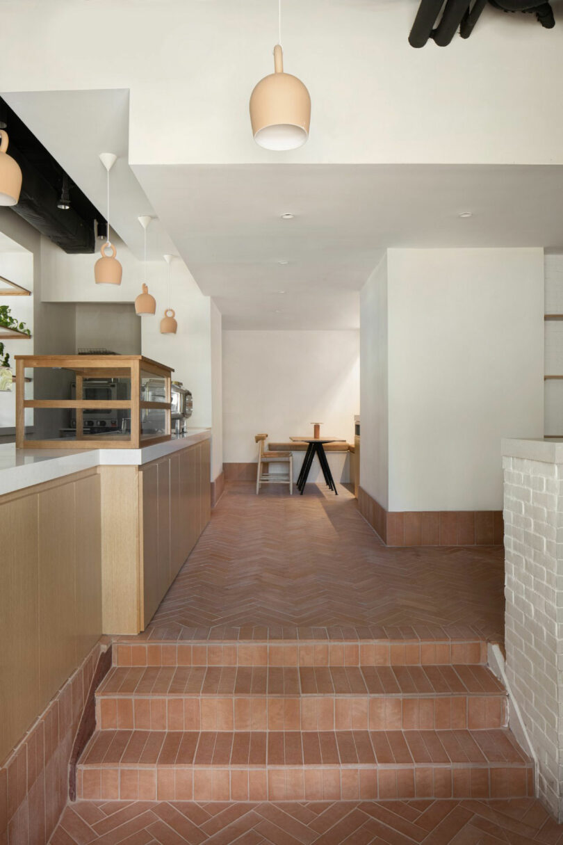 A minimalist cafe interior by Nicki Gitlin features terracotta tile flooring, light wood cabinetry, pendant lights, and a small dining area with a table and chairs in the background.