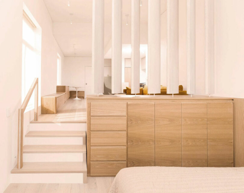 Minimalist bedroom by Nicki Gitlin featuring light wood cabinetry, vertical white slats as a room divider, and steps leading to a raised sitting area bathed in natural light.
