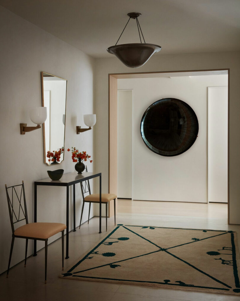 A minimalist hallway by Nicki Gitlin features a console table, two chairs, a geometric-patterned rug, wall sconces, and a large round mirror on the far wall.