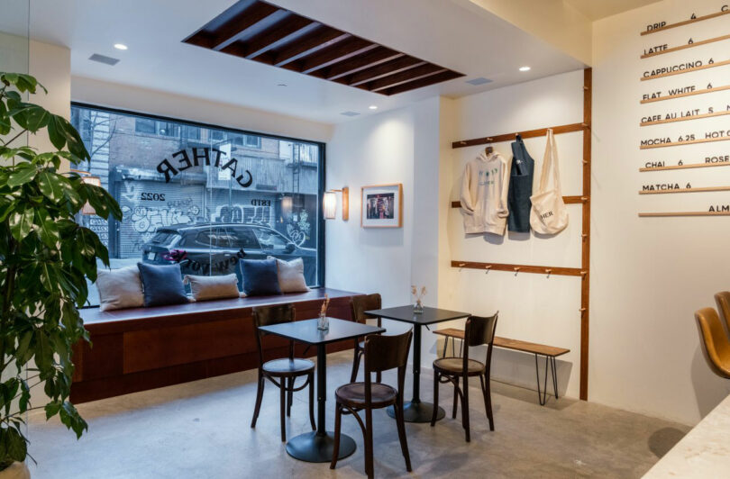A modern cafe interior by Nicki Gitlin with wooden tables and chairs, a bench by a large window, coat hooks with bags, wall art, and a potted plant. A street and parked cars are visible outside.