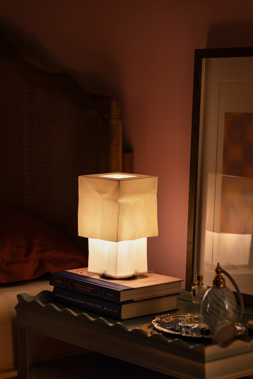 A square-shaped table lamp with a soft, white shade sits on a stack of books next to a framed picture, perfume bottles, and a decorative tray on a bedside table.