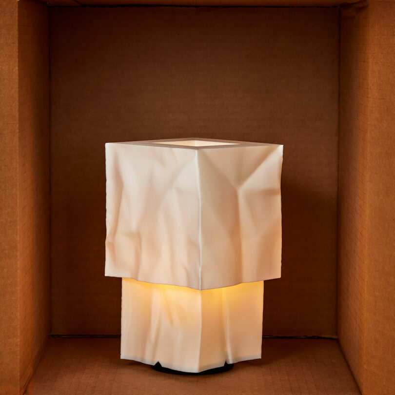 A square, white, wrinkled lampshade illuminated from within, placed inside a plain brown cardboard box.