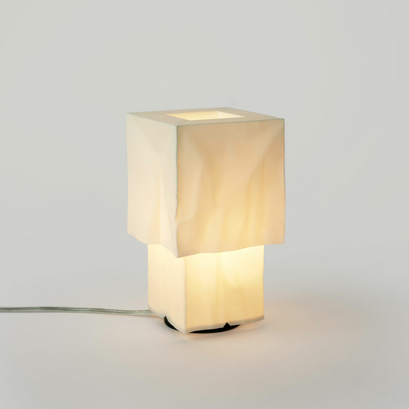 A modern table lamp with a white, crumpled square-shaped shade, emitting a soft warm light, and a visible power cord on a plain background.