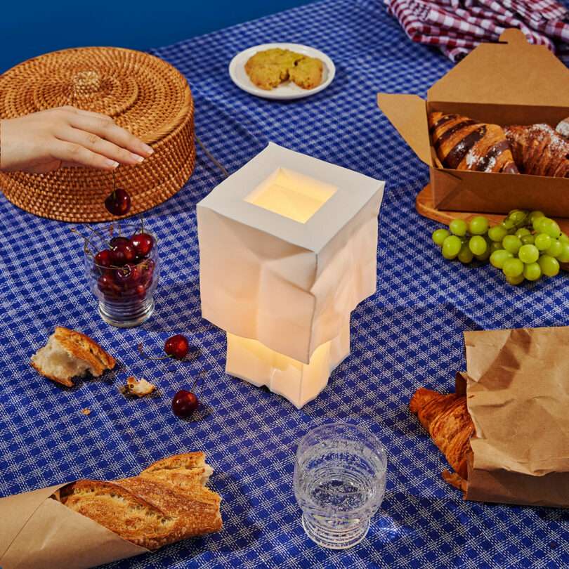 A picnic table with a patterned blue cloth, various breads, grapes, cherries, pastries, a glowing geometric lantern, and a hand reaching for a round wicker basket.