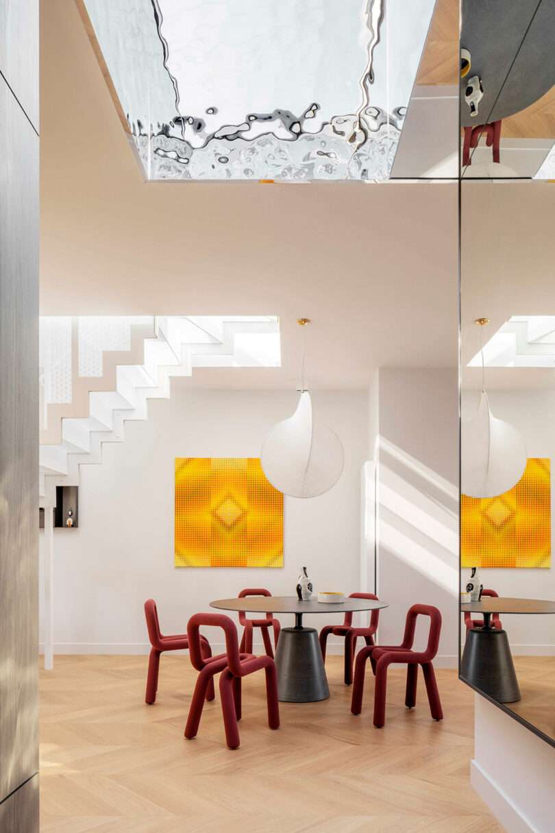 Modern dining area with red chairs, round tables, geometric yellow wall art, a large circular light fixture, and a reflective ceiling panel above.