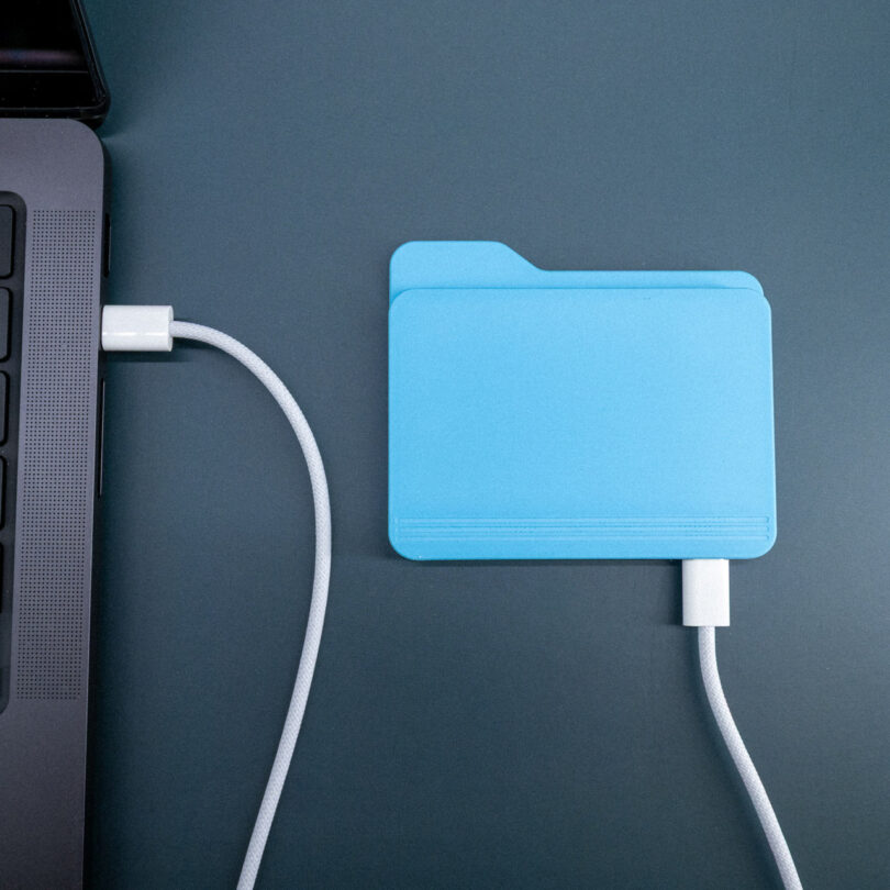A blue folder-shaped external drive is connected to a laptop with a white USB cable on a dark surface.