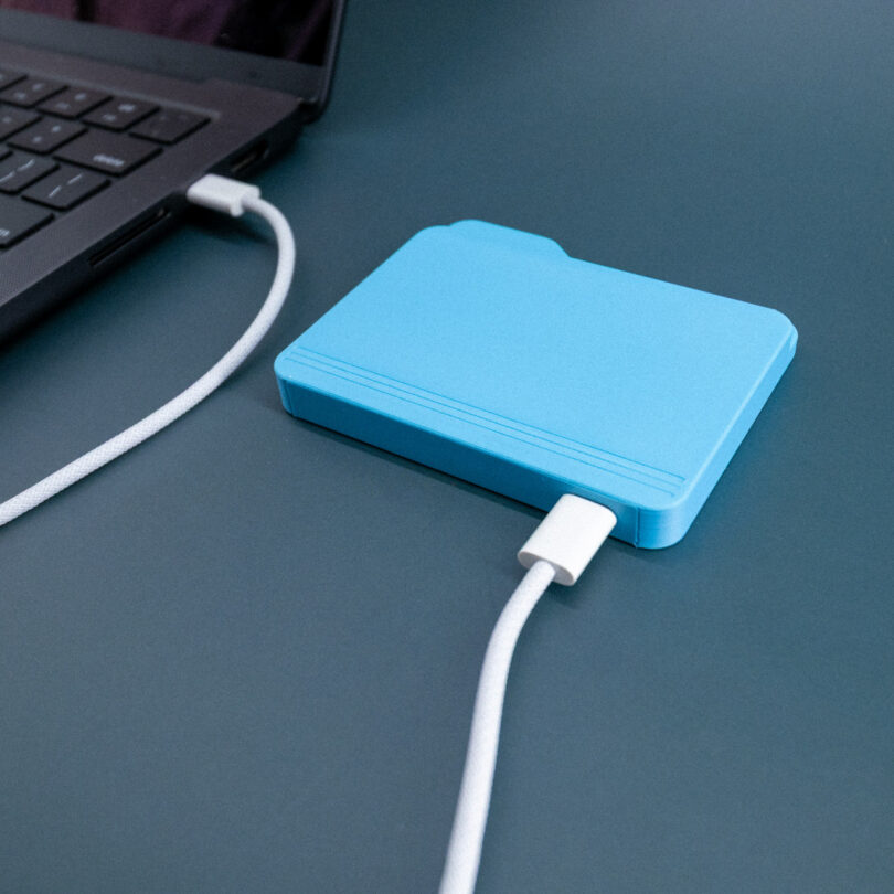 A blue external hard drive shaped like a folder is connected to a laptop with a white cable on a blue surface.