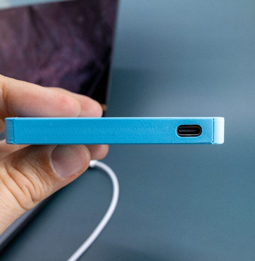 A hand holds a blue device with a USB-C port visible on the edge, and a white cable is partially shown in the background.