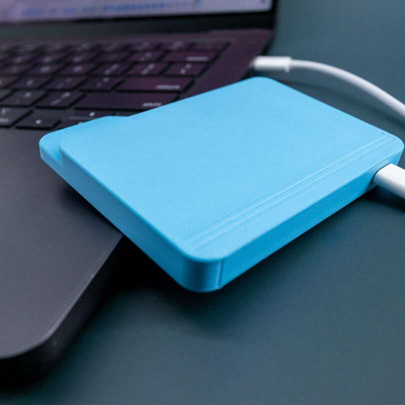 A blue external hard drive connected to a laptop via a white cable, placed on a dark surface.