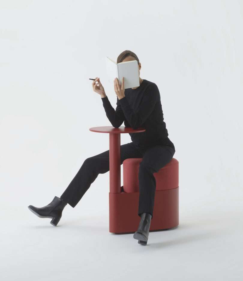 Person dressed in black sits on a round red ottoman beside a matching side table, holding a book and a pen, with a minimal white background