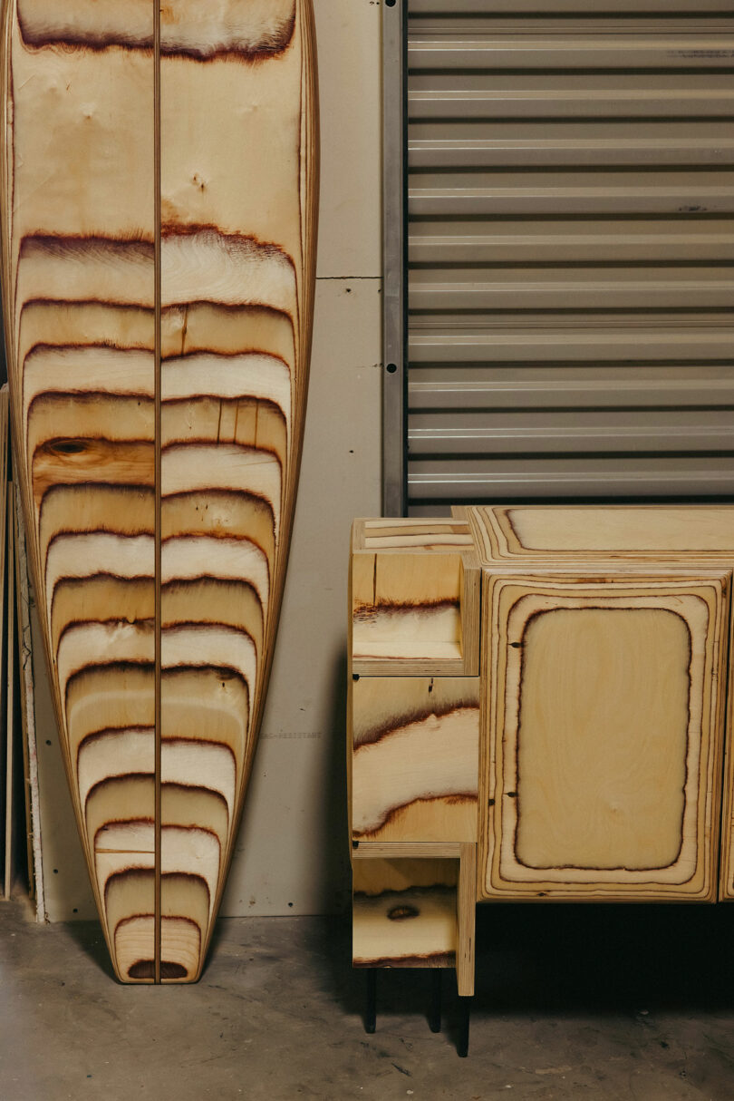 A wooden surfboard and a wooden cabinet with matching natural burn patterns, reminiscent of Kelly Wearstler’s signature style, stand beside a metal garage door in an indoor setting.