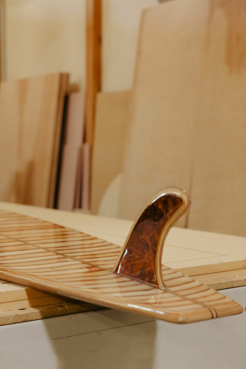 A close-up of a wooden surfboard with a varnished wooden fin, resting on a workbench in a workshop with plywood sheets in the background, exuding a style reminiscent of Kelly Wearstler’s distinctive aesthetic.