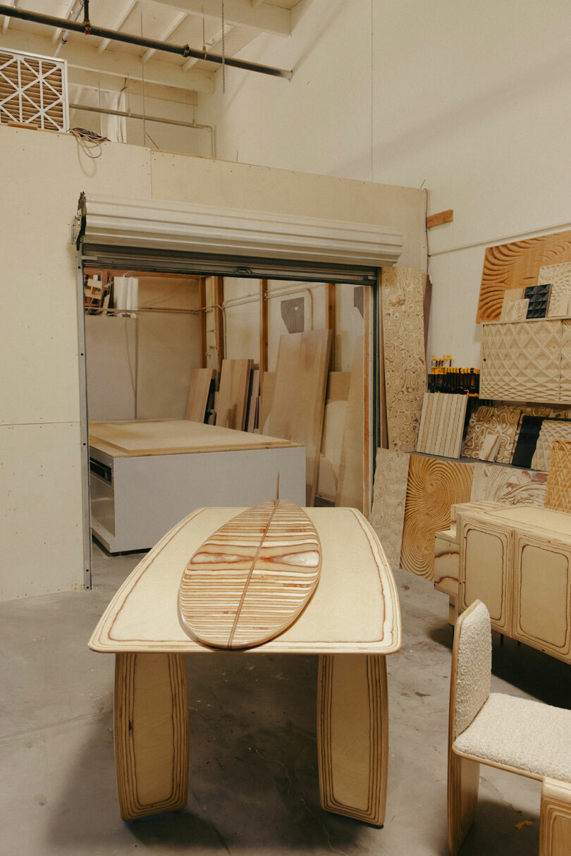 A surfboard with a wood grain pattern rests on a matching wooden table in a workshop filled with plywood sheets and unfinished furniture, echoing the sophisticated style of Kelly Wearstler.