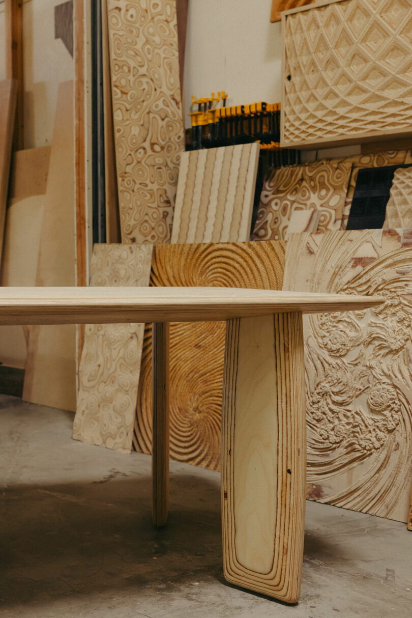 A wooden table stands in a workshop, surrounded by various intricately carved wooden panels, showcasing a style reminiscent of Kelly Wearstler's bold and artistic designs.