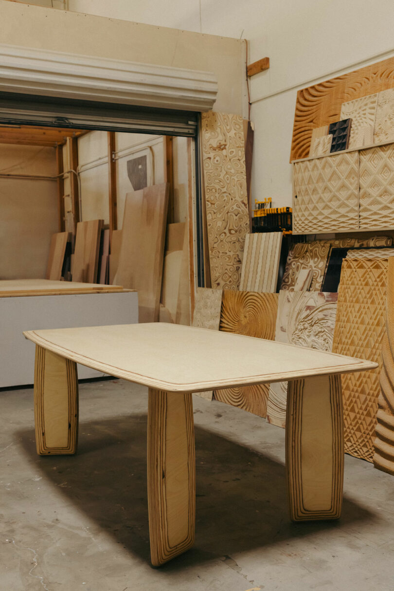 A wooden table with thick, curved legs stands in a workshop, surrounded by sheets of patterned wood and woodworking tools, exuding a style reminiscent of Kelly Wearstler's bold and unique designs.