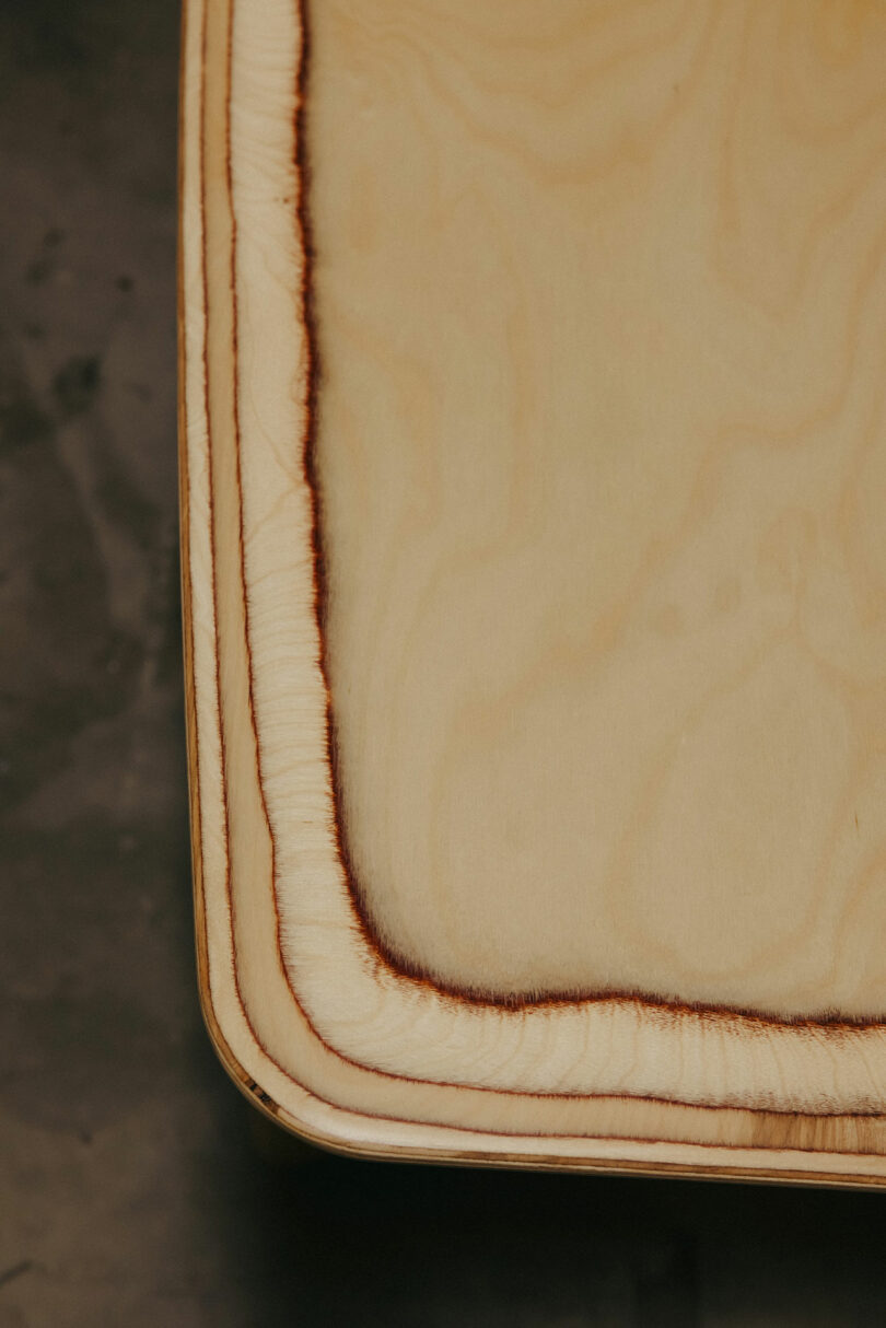 Close-up of a light wooden surface with a rounded edge inspired by Kelly Wearstler, featuring a distinct dark burn mark outlining the border on a neutral background.