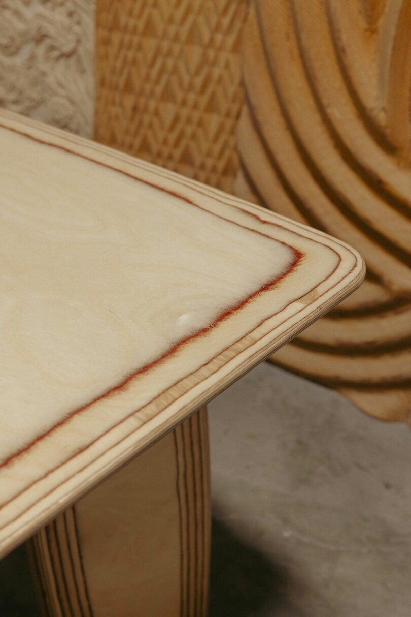 Close-up of a plywood table corner with visible layered edges, showing smooth surface and natural wood grain. In the background, carved wooden objects evoke Kelly Wearstler’s modern design sensibility.