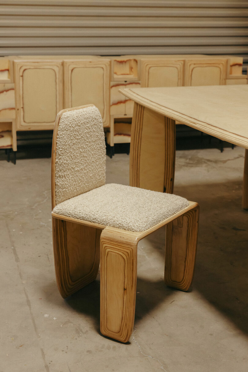 A wooden chair with a curved design and cream textured cushions, inspired by Kelly Wearstler, is positioned near a matching wooden table in an indoor setting.