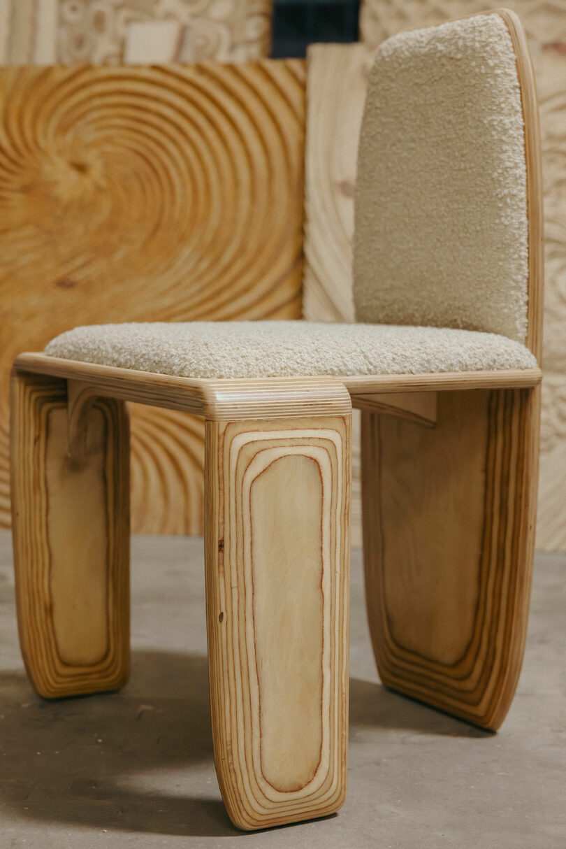 A modern chair with curved, layered wood legs and a cream-colored upholstered seat and backrest, inspired by Kelly Wearstler, set against a background of unfinished wooden panels.