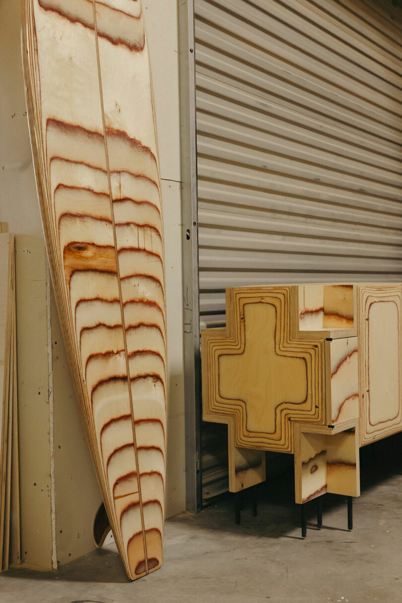 Sheets of plywood with visible wood grain lean against a wall, alongside two wooden structures with cross-shaped cutouts on black metal legs—evoking a workshop scene with a hint of Kelly Wearstler-inspired style.
