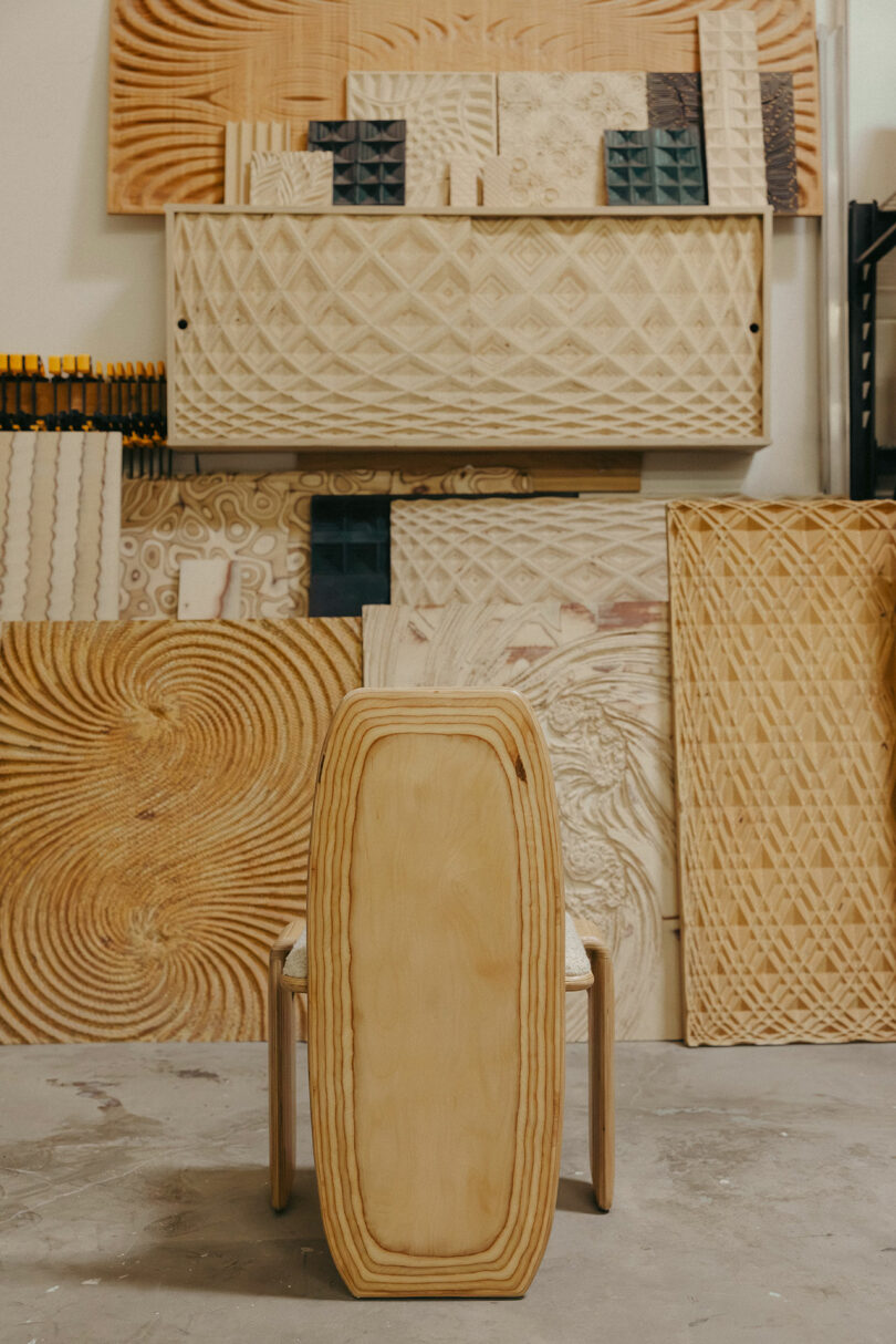 A wooden chair sits before multiple carved and patterned wooden panels, evoking a Kelly Wearstler-inspired aesthetic, in a sunlit workshop.