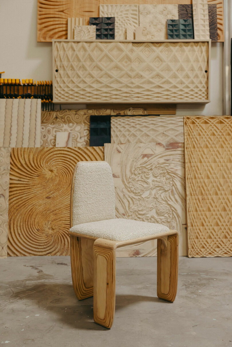 A modern wooden chair with a cushioned seat and backrest, inspired by Kelly Wearstler’s bold designs, sits in front of decorative carved wood panels in a stylish workshop setting.
