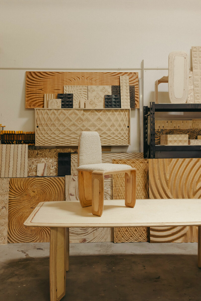 A modern wooden chair with a cushioned seat is displayed on a wooden table, complemented by carved wooden panels arranged on shelves and against the wall, evoking a Kelly Wearstler-inspired aesthetic.