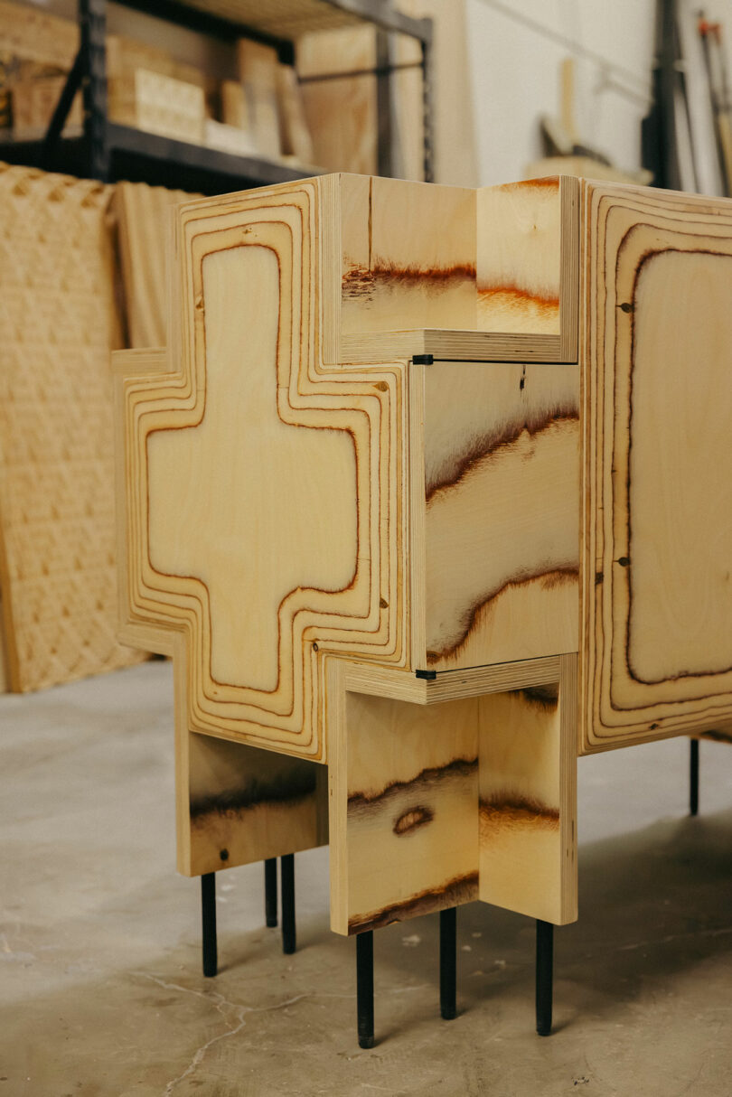 A wooden cabinet with a geometric, cross-shaped design and black metal legs, reminiscent of Kelly Wearstler’s bold style, stands in a workshop setting with tools and wood in the background.
