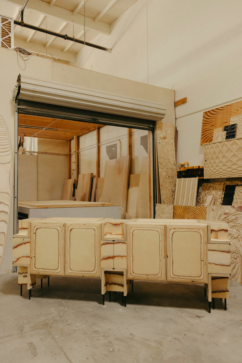 Wooden cabinets in various stages of construction stand in front of a partially open garage door, bringing to mind Kelly Wearstler’s artful approach to craftsmanship in a woodworking shop filled with wood panels and tools.