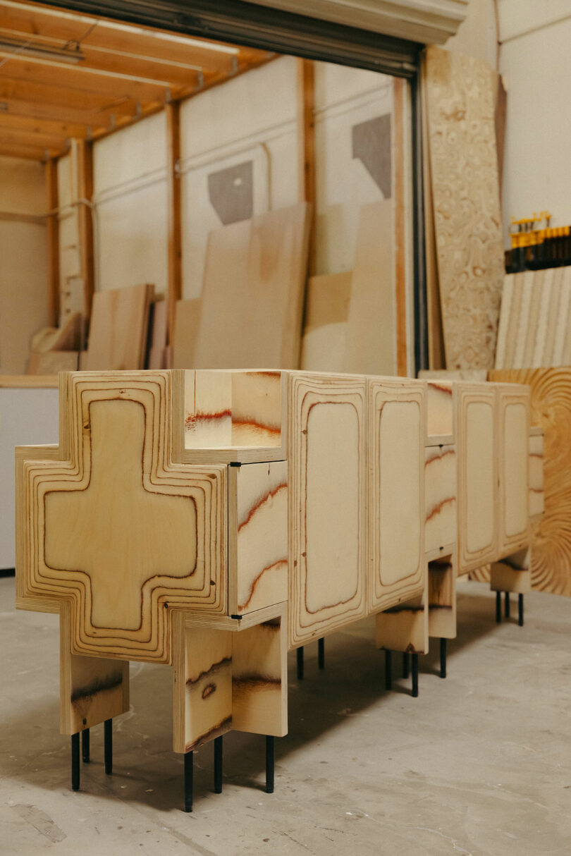 A wooden cabinet with cross-shaped ends and layered plywood design, inspired by Kelly Wearstler’s style, stands on black metal legs in a woodworking shop.