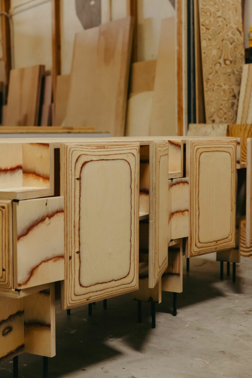 Several unfinished wooden cabinets with visible grain patterns stand on black legs in a workshop, evoking a raw aesthetic reminiscent of Kelly Wearstler, surrounded by plywood sheets and woodworking materials.