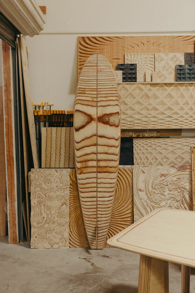 A wooden surfboard with a symmetrical grain pattern stands upright against a wall, surrounded by various plywood sheets featuring intricate patterns in a workshop reminiscent of Kelly Wearstler’s bold style.