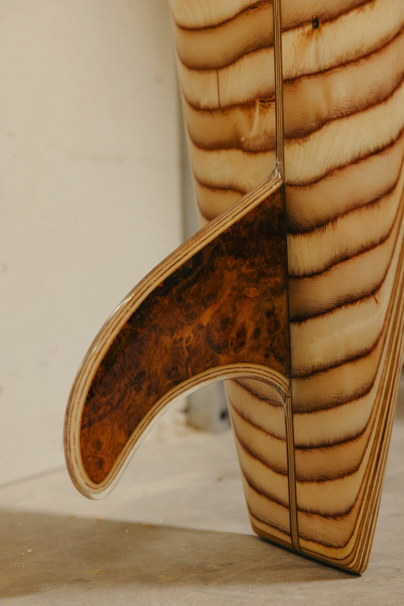 A close-up of a wooden surfboard tail and fin, featuring layered wood grain and a contrasting, darker wood inlay on the fin—evoking the signature style and sophistication of Kelly Wearstler.