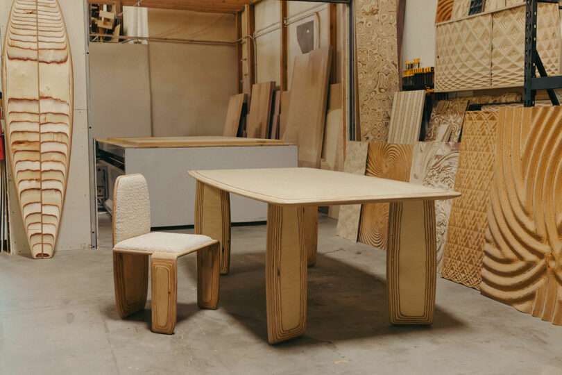 A wooden chair and table with layered edges, inspired by Kelly Wearstler’s bold style, are placed in a workshop, surrounded by various wood panels and tools.