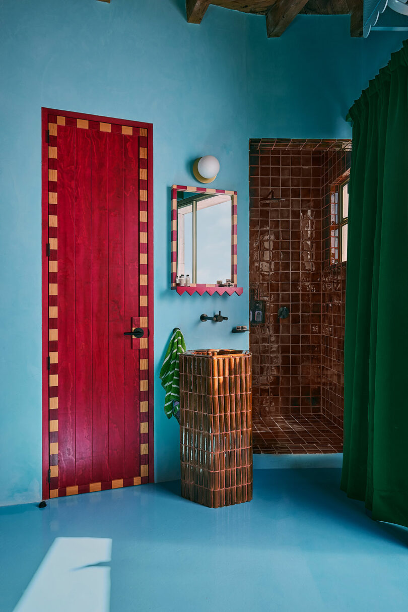 A bathroom with a red door, blue walls, brown tiled shower, green curtain, wicker pedestal sink, and a red mirror above the sink.