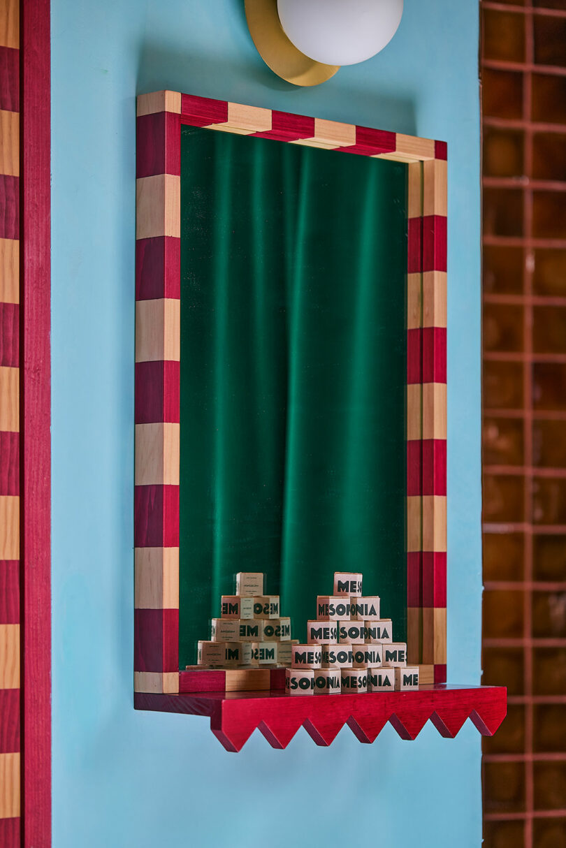 Rectangular mirror with a red and beige checkered frame reflecting wooden blocks on the shelf spelling 