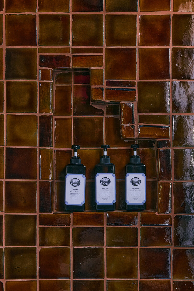 Three dark pump bottles with white labels mounted on a brown tiled wall, arranged vertically in a recessed tile section.