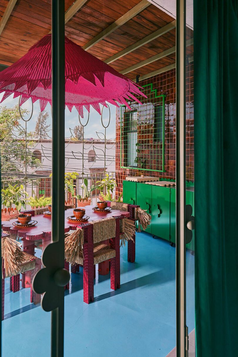 Colorful outdoor dining area with a pink umbrella, straw-trimmed chairs, green cabinets, and a blue floor, viewed through glass doors.
