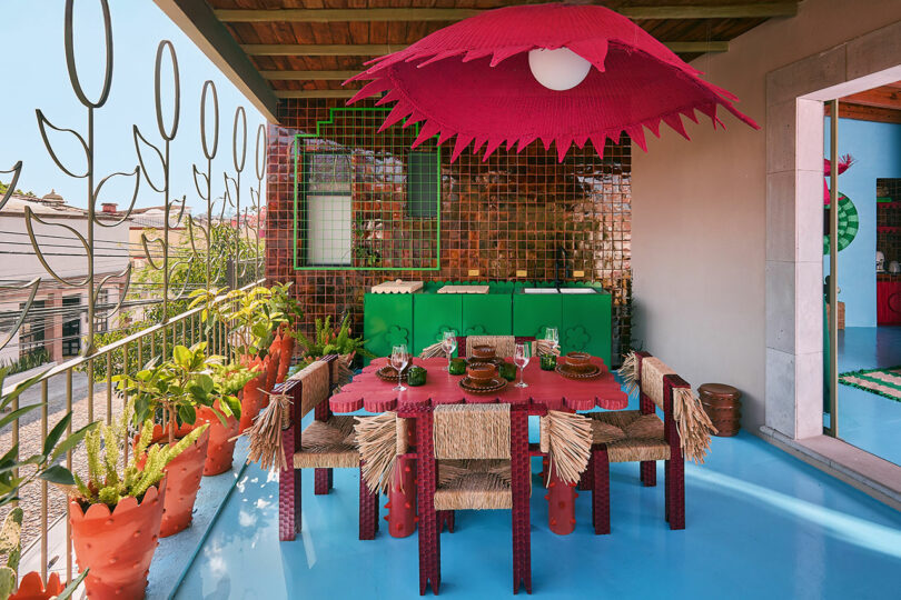 Colorful outdoor dining area with a pink umbrella, straw-trimmed chairs, terracotta planters, mosaic tiles, and green cabinets on a blue floor.