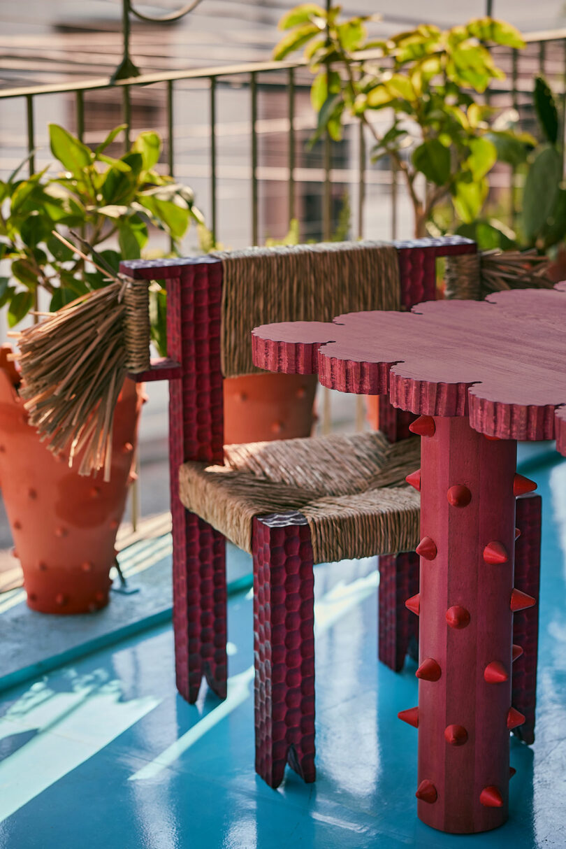 A unique red table with textured legs and a matching chair with woven seat and backrest sits on a blue floor, surrounded by potted plants on a balcony.