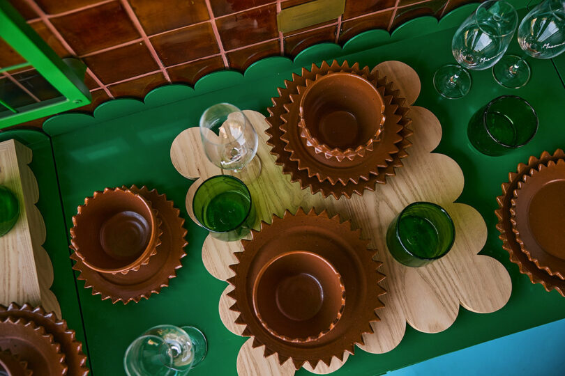 Table set with brown ceramic bowls, green glass cups, wine glasses, and wooden placemats on a green surface, with brown tiled backsplash in the background.