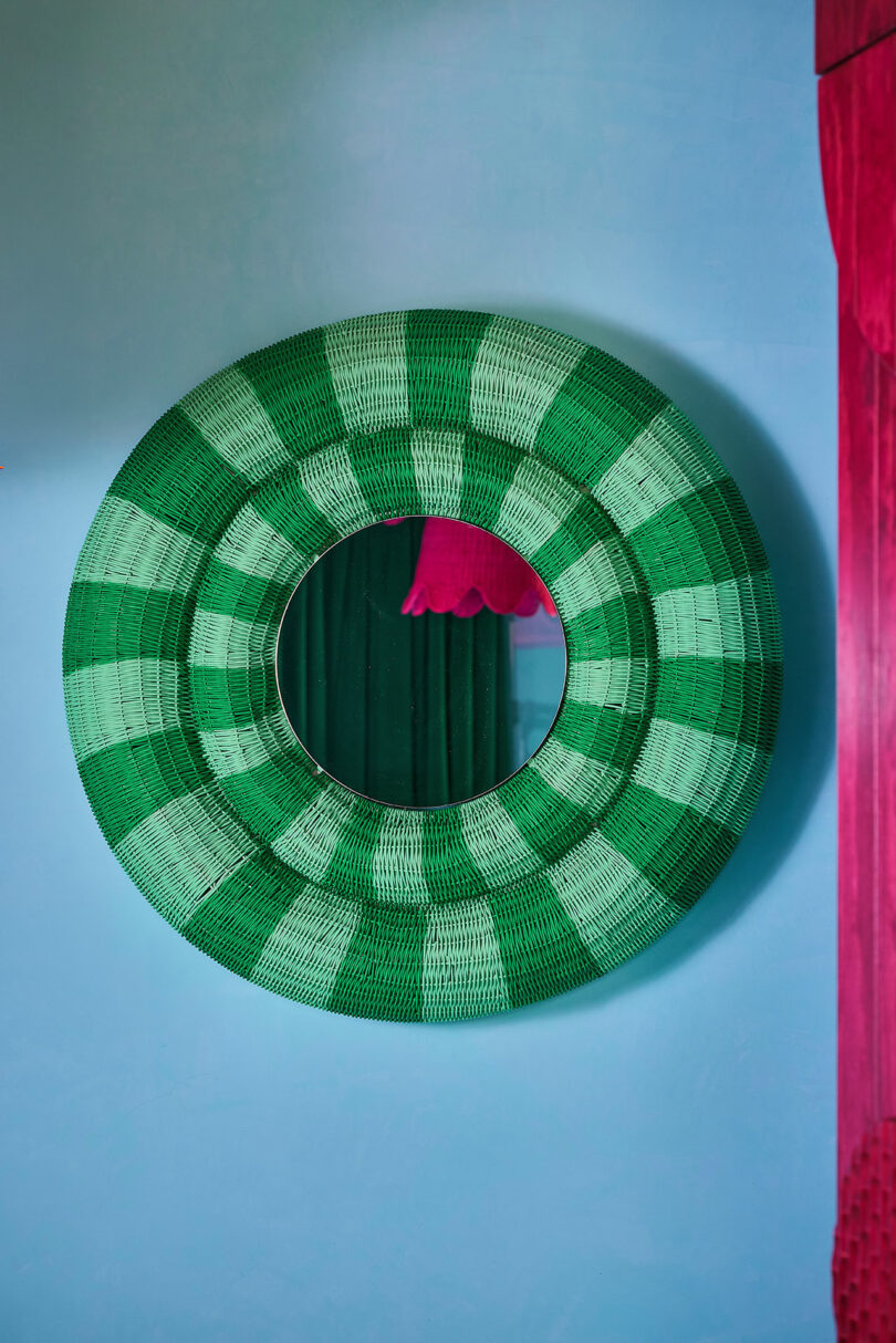 A round mirror with a green checkered woven frame hangs on a light blue wall with pink and green curtains partially visible in the reflection.